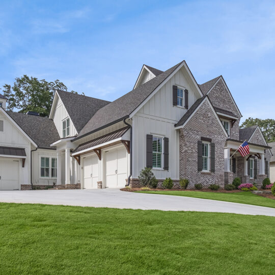 A large, modern house with a combination of brick and white siding exterior, featuring multiple dormer windows and gabled roofs. The driveway leads to an attached garage. A well-manicured lawn and American flag complete the scene.