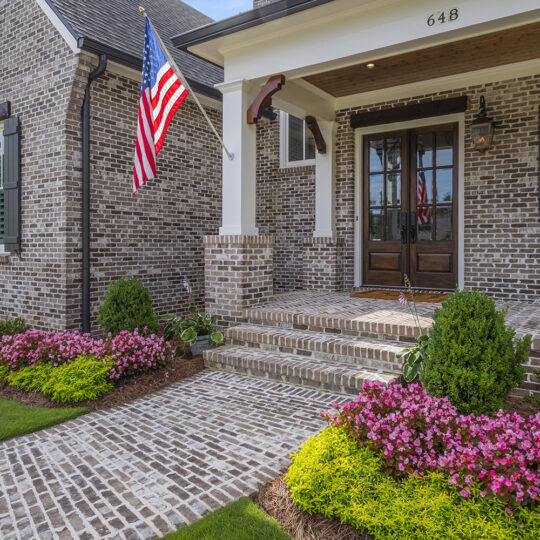 A brick house with a porch, an American flag, and black shutters. The front yard features vibrant pink flowers, manicured bushes, and a neatly trimmed lawn. Steps lead up to a dark wooden door, and the house number 648 is visible above the entrance.