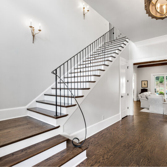 A modern interior features a staircase with dark wooden steps and a black railing. The space includes a seating area with light furniture, a large window, and a view of an outdoor patio. Wall sconces and a ceiling light provide illumination.