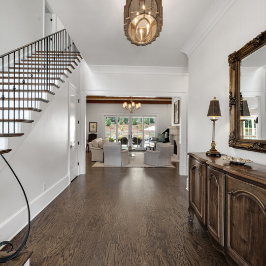 A spacious, elegant entryway with dark hardwood floors and a staircase with black railings on the left. A wooden sideboard with lamps and a large mirror is on the right. In the background, a living area with sofas and large windows is visible.