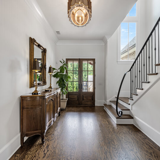 A bright hallway with wooden flooring and a staircase to the right. A wooden sideboard with a decorative mirror and lamp is on the left. Large double doors with glass panes open to the outside. A potted plant sits near the door, and a chandelier hangs above.