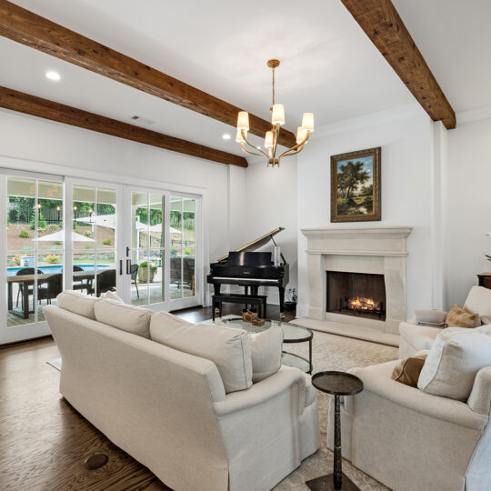 A cozy living room features beige sofas around a glass coffee table, facing a fireplace with a painting above. A black grand piano sits nearby. Wooden beams adorn the ceiling, and large windows offer a view of a garden and pool outside.