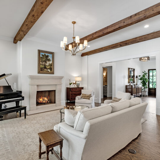 Living room with a grand piano, beige sofas, a wooden coffee table, and a lit fireplace under a painting. Exposed wooden ceiling beams and a chandelier add charm. Hardwood floors and a doorway to another room complete the setting.