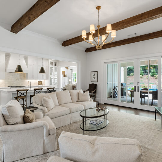 Spacious living room with white sofas and armchairs, a round coffee table, and a grand piano by the window. Exposed wooden beams and a chandelier enhance the room. Large windows reveal a view of an outdoor patio and greenery.