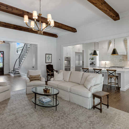 Spacious living room with a beige sofa and armchairs on a patterned rug, open to a modern white kitchen with an island and bar stools. Exposed wooden beams on the ceiling and a chandelier add warmth. A staircase is visible in the background.