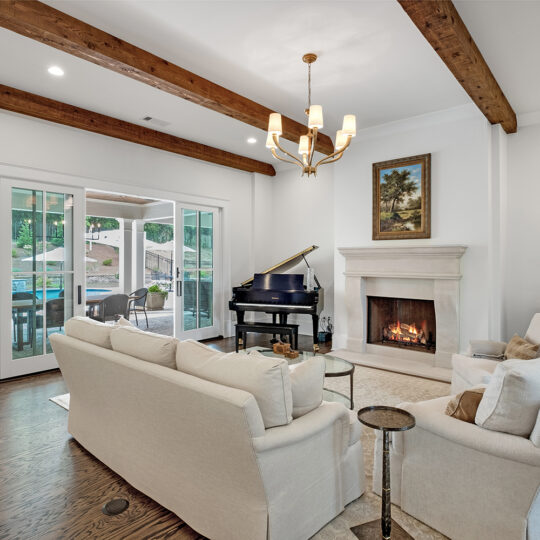 A cozy living room with white sofas, a wooden coffee table, and a lit fireplace. A grand piano is near sliding glass doors leading to an outdoor patio. The room features wooden ceiling beams, art on the walls, and a chandelier.