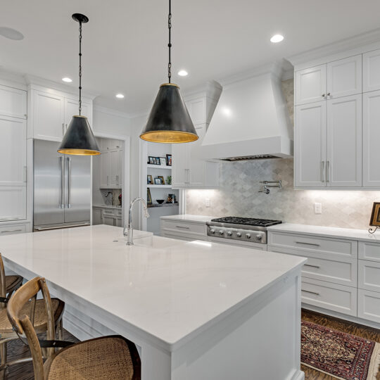 A modern kitchen with white cabinetry, a large island with a marble countertop, and wicker stools. The space includes stainless steel appliances, two black pendant lights, and a decorative rug on wooden flooring.