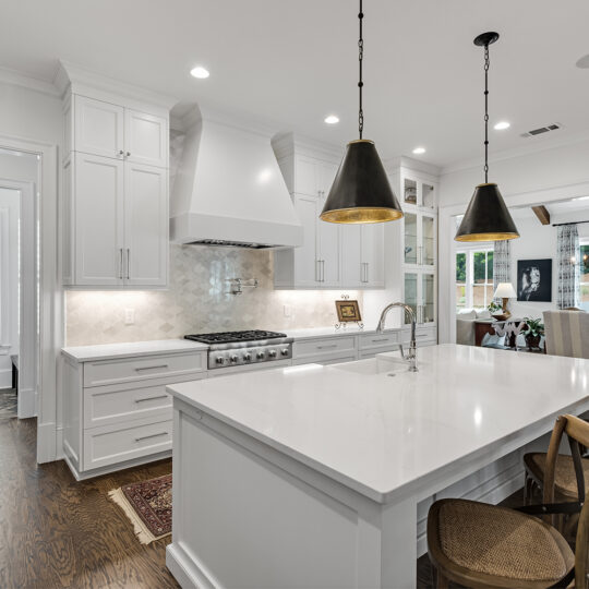 A modern kitchen featuring white cabinetry, a large island with a white countertop, wooden bar stools, pendant lighting, and stainless steel appliances. The open layout leads to a dining area and a room with large windows.