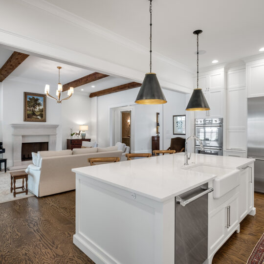 Open-concept kitchen and living area with a white island and farmhouse sink, stainless steel appliances, wooden floor, and pendant lights. The living room features a grand piano, fireplace, beams on the ceiling, and neutral-toned furniture.