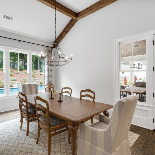 Dining room with a wooden table and six chairs, a large window showing an outdoor pool, and a chandelier overhead. Neutral tones with wooden beams on a high ceiling. Glass doors lead to a sunroom with more seating and a view of the yard.