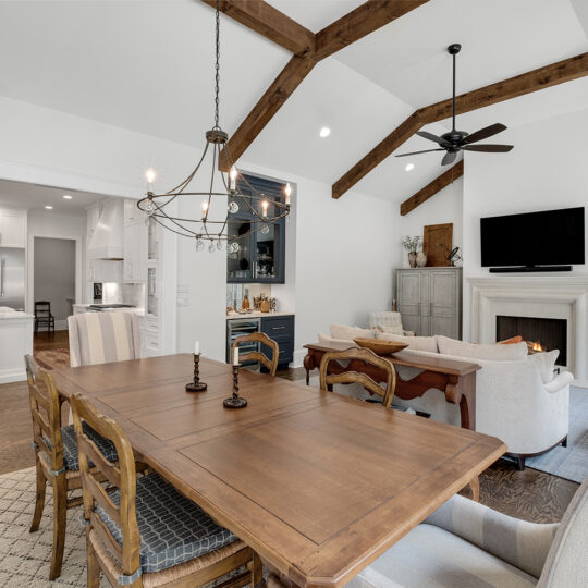 Open-concept living room with vaulted ceiling and wood beams. Features include a dining table, chandelier, beige sofas facing a fireplace with a TV above, and a kitchen with white cabinets visible in the background. Neutral tones throughout.