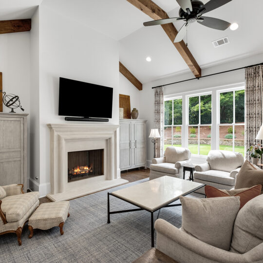 A cozy living room with a high ceiling features a stone fireplace below a wall-mounted TV. Surrounding the space are a beige couch, armchairs, a coffee table, and a fainting couch near large windows with curtains. Wooden beams accent the ceiling.