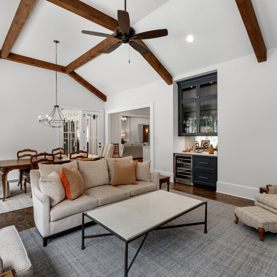 Spacious living room with high vaulted ceiling featuring wooden beams, a beige sofa, armchairs, and a coffee table. A dining area with a chandelier is visible in the background. A built-in shelving unit is on one wall. Neutral tones dominate the decor.