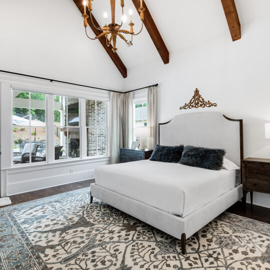 Spacious bedroom with a vaulted ceiling and exposed wooden beams. Theres a bed with a tall headboard and dark pillows, two dark nightstands with a lamp and framed photos, a large ornate rug, and a view of a patio and pool through glass doors.