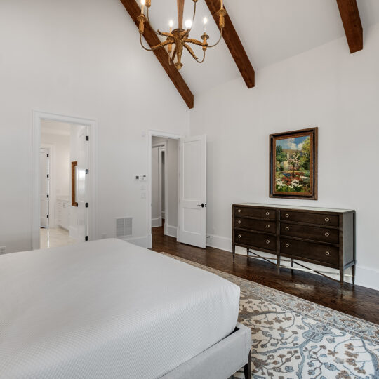 A bedroom with vaulted ceilings and wooden beams. Theres a neatly made bed with gray pillows, a dark wood dresser with a painting above it, and a patterned rug on the floor. A chandelier hangs from the ceiling, and an open door reveals a hallway.