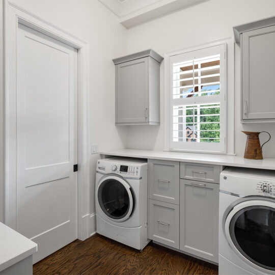 Laundry room with a white and gray color scheme. It features a front-loading washer and dryer side by side, gray cabinets, a countertop, and a window with shutters. A brown pitcher sits on the countertop above the dryer.