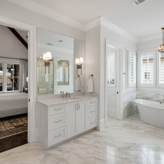 Spacious bathroom with white marble floor, a freestanding bathtub, and a double-sink vanity with mirrors. It features a modern chandelier and opens to a bedroom with a large window and a bed.