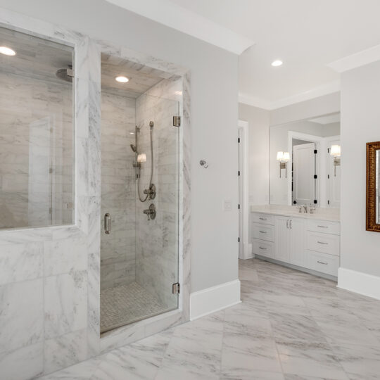 Luxurious bathroom with a large marble-tiled shower featuring glass doors. The room also has a double vanity with cabinets, a wide mirror, wall-mounted lights, and a framed mirror. The floor is also covered in marble tile, enhancing the elegant design.