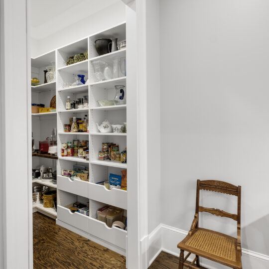 A neatly organized pantry with white shelves containing jars, cans, and various kitchen items. The space has hardwood floors and white walls. A wooden chair with a woven seat is placed against the wall near the pantry entrance.