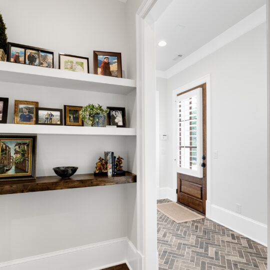 Entrance hallway with light walls and wooden interiors. Photos and small decorations are on shelves to the left. The floor features a patterned tile leading to a wooden door with glass panels. Natural light fills the space.