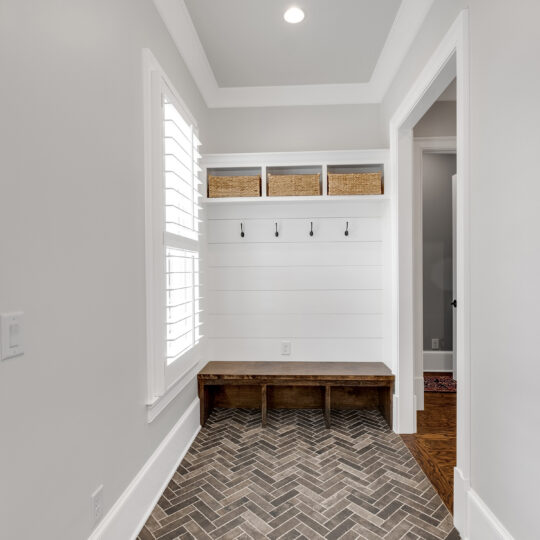 A mudroom with a wooden bench and herringbone-patterned tile floor. Above the bench are four black hooks and three wicker baskets. The walls are white, and a window on the left allows natural light in.