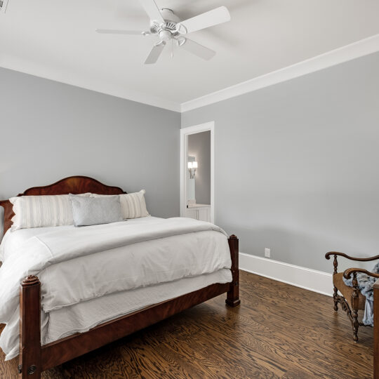 A bedroom with a large wooden bed featuring white bedding against a gray wall. A bedside table with a lamp is on the left, a wooden chair with a blanket is on the right, and theres a ceiling fan above. The floor is hardwood.