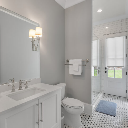 A bright bathroom with a white vanity and sink, a large wall mirror, and a light fixture above. Theres a toilet beside the vanity. A glass-door shower is at the back, and a windowed door leads outside. The floor has a black and white hexagonal tile pattern.