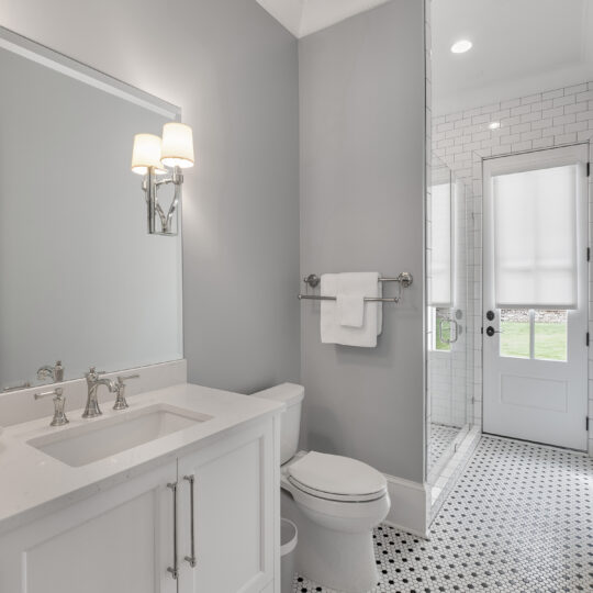 A modern bathroom with light gray walls and a white vanity. It features a large mirror, a toilet, and a glass-enclosed shower. The floor has black and white hexagonal tiles, and theres a door with a frosted window. A wall sconce provides lighting.