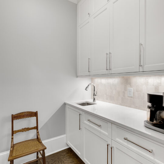 A modern kitchen with white cabinets and a white countertop. There is a sink with a faucet and a coffee maker on the counter. A wooden chair with a woven seat sits on the hardwood floor against a light gray wall.