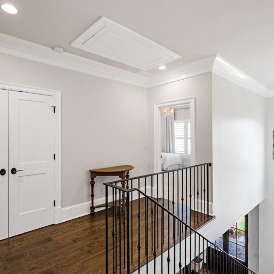 A bright hallway features wooden flooring and white walls. A dark metal railing lines the stairs, leading to a door. A small wooden table sits against the wall, and sunlight streams through tall windows, providing a view outside.