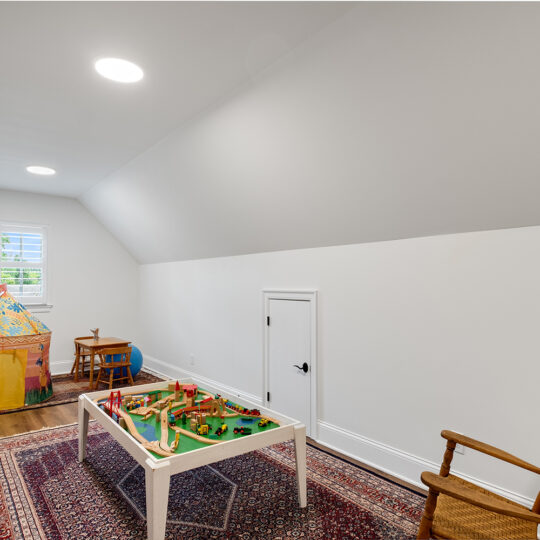 A playroom with a sloped ceiling, featuring a small table with toys, a play tent, a wooden rocking chair, and a colorful rug on the wooden floor. A small door is set into the wall, and two round ceiling lights illuminate the room.