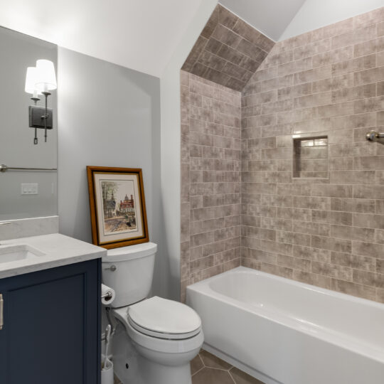 A modern bathroom featuring a bathtub with gray tiled walls, a toilet next to a blue vanity with a countertop and a sink, a decorative painting leaning on the toilet, and an overhead light fixture. The floor has hexagonal tiles.