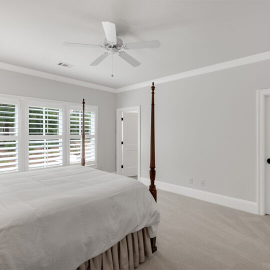 A bright bedroom with light gray walls and a beige carpet. It features a bed with white linens and a four-poster frame, a window with white shutters, a ceiling fan, and two closed doors. Natural light streams in from the window.