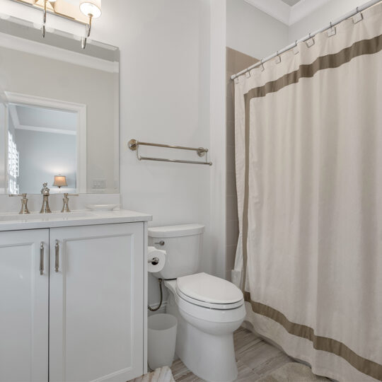 A modern bathroom with a white vanity and mirror on the left, a toilet in the center, and a shower with a beige curtain on the right. The room features light-colored walls and flooring, with a towel rack and overhead light fixture.