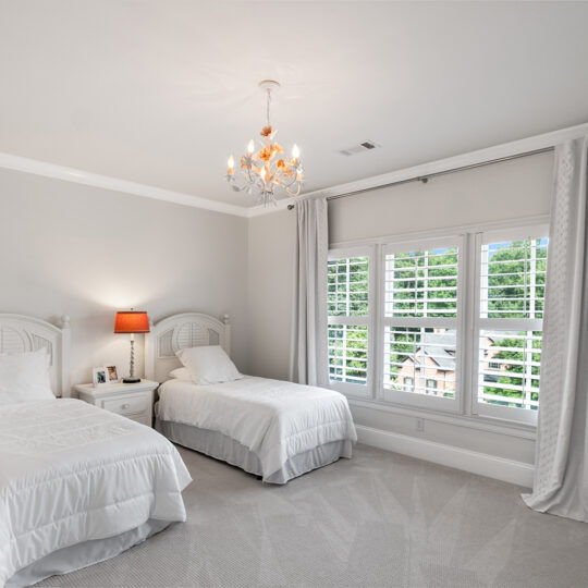 A bedroom with two twin beds featuring white bedding and headboards. A red lamp sits on a nightstand between them. A window with drapes and blinds lets in sunlight. A chandelier hangs from the ceiling. Beige carpet covers the floor.