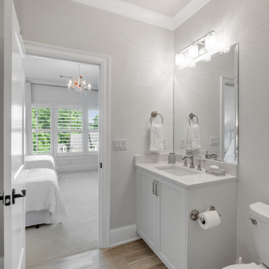 Modern bathroom with a white vanity, mirror, and bright lighting. Theres a toilet and a toilet paper holder with a roll. The room leads to a bright bedroom with a bed, windows, and a chandelier. Wood flooring in the bathroom transitions to carpet in the bedroom.