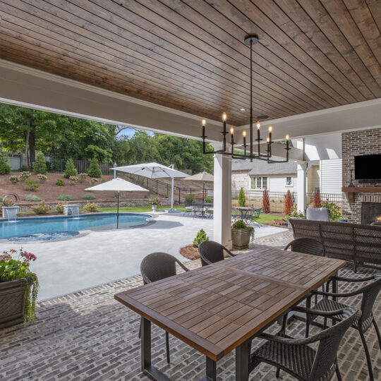 Covered patio with wooden ceiling and chandelier, overlooking a backyard pool. Patio features a long dining table, chairs, grill, and fireplace with a TV above. Pool area has lounge chairs, umbrellas, and is bordered by landscaped greenery.