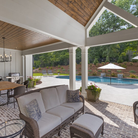 Outdoor patio with wicker furniture, including a sofa, chairs, and a table, overlooks a pool. The ceiling is wooden with a chandelier. The pool area includes umbrellas and lounge chairs, surrounded by greenery and trees under a clear sky.