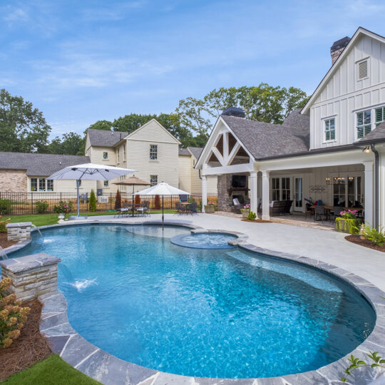 A luxurious backyard features a large swimming pool with a small attached jacuzzi. Surrounding the pool are lounge chairs, umbrellas, and lush landscaping. A white, two-story house with a spacious covered patio sits to the right.