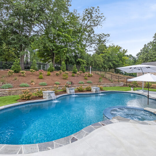 A backyard with a clear swimming pool featuring a stone border. The pool has three cascading water features and a small attached hot tub. It is surrounded by a patio, large umbrella, and lush greenery on a landscaped hill.