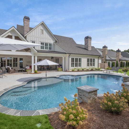 Spacious backyard with a modern house, featuring a large, curvy swimming pool surrounded by landscaped plants. Theres patio furniture under umbrellas and a lawn area, all beneath a clear blue sky.