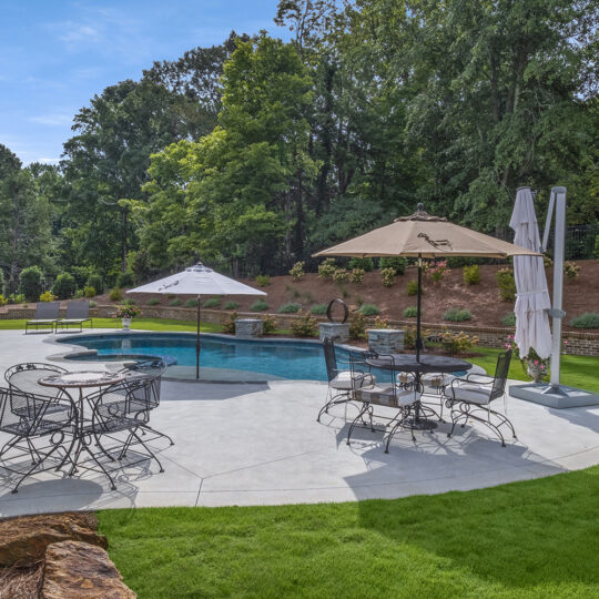 A backyard with a kidney-shaped swimming pool surrounded by concrete patio. Several lounge chairs and tables with umbrellas are placed around the pool. Green grass, trees, and shrubs border the area under a clear blue sky.