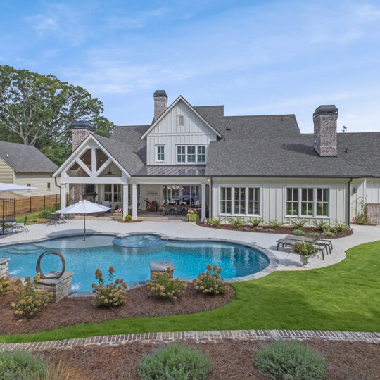 A spacious backyard features a modern house with a gable roof. It includes a circular pool, hot tub, white poolside umbrellas, lounge chairs, and a manicured lawn. A wooden fence and neighboring houses are visible under a partly cloudy sky.