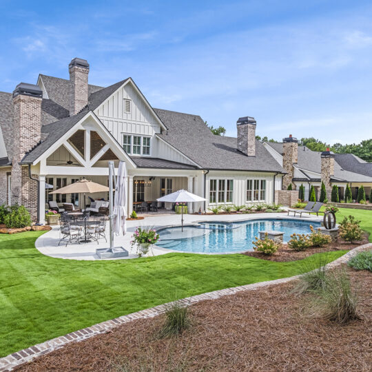 A spacious backyard features a large, modern house with stone chimneys and a gabled roof. A kidney-shaped pool is surrounded by neatly manicured lawn, patio furniture, and lush greenery under a clear blue sky.