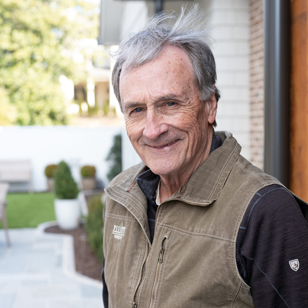 Elderly man with gray hair smiles outdoors, wearing a brown vest over a black shirt. Hes in a garden with green plants, trees, and a white wall in the background.