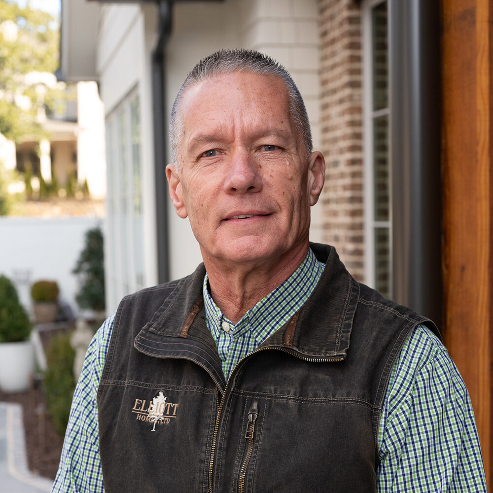 A man with short gray hair is wearing a dark vest over a green plaid shirt. He stands outside next to a brick wall, with a house and shrubs in the background.