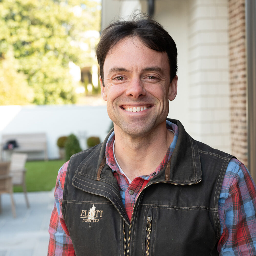 A smiling man with dark hair is wearing a plaid shirt and a vest with a logo. He stands outdoors in a sunlit area with a building and greenery in the background.