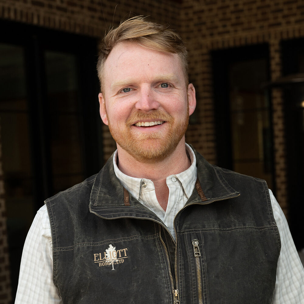 A man with short, light hair and a beard is smiling. He wears a checkered shirt under a black vest with the text Elliott Homes LTD embroidered on it. The background features a brick wall.